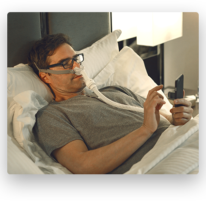 A male in bed using his phone with an AirMini nose mask in place